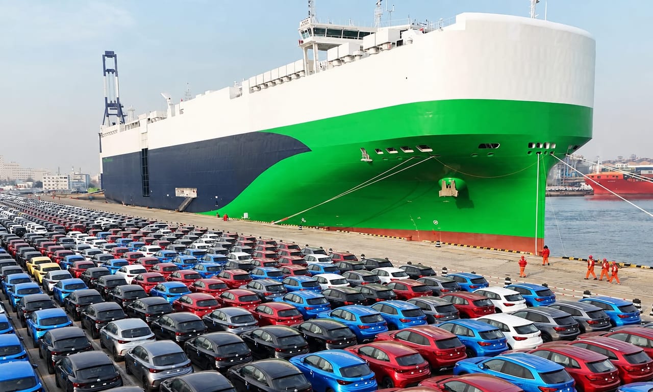 Rows of new cars lined up at a port vehicle terminal beside a RoRo car-carrier vessel at dusk